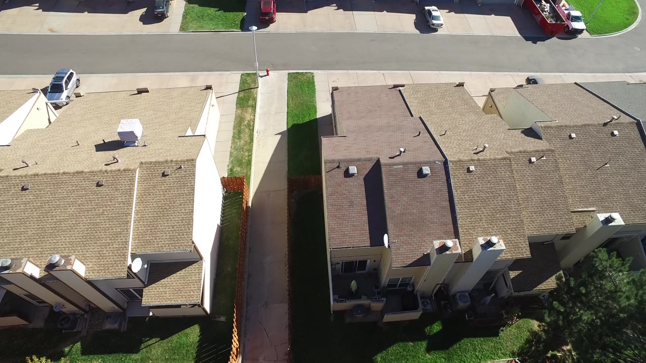 Overhead drone frame in Hidden Lake, Colorado showing tan and brown house roofs, a straight street, lawns, driveways, parked cars, and no visible people.