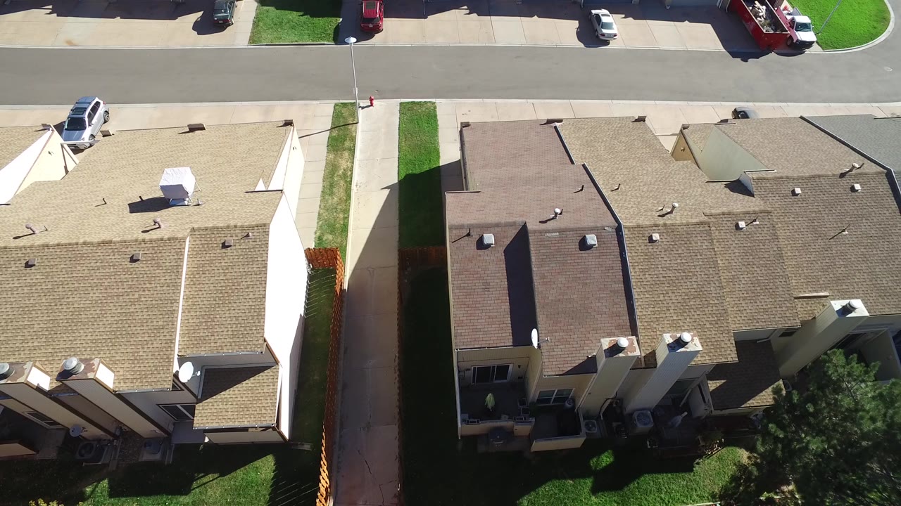 High overhead view in Hidden Lake, Colorado with attached rooftops, a pale driveway, narrow grass strips, a white parked vehicle near the curb, and no visible people.