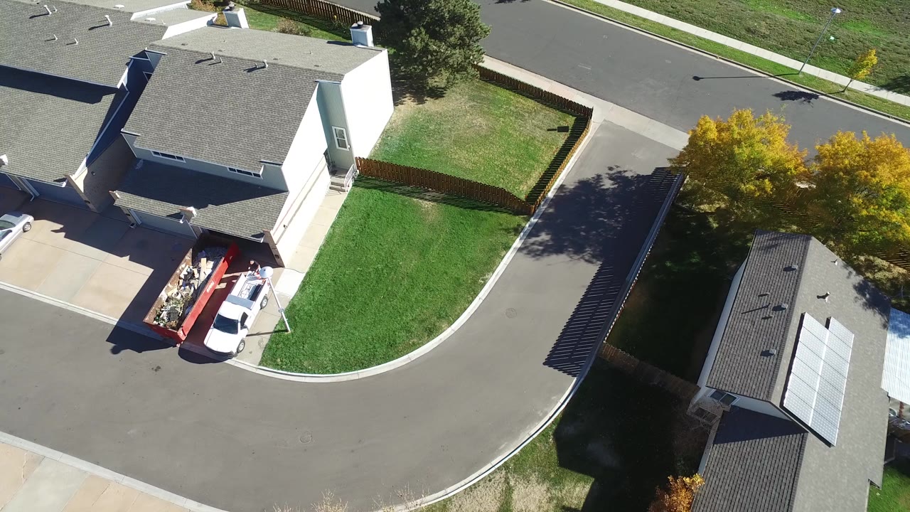 High oblique aerial frame over Hidden Lake, Colorado, showing private homes, curved streets, green lawns, a white vehicle with red trailer, and yellow autumn trees.