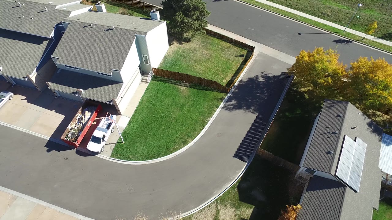 Aerial view in Hidden Lake, Colorado, with a white vehicle and red trailer near a driveway, fenced yards, green lawns, rooftops, and yellow trees along the street.