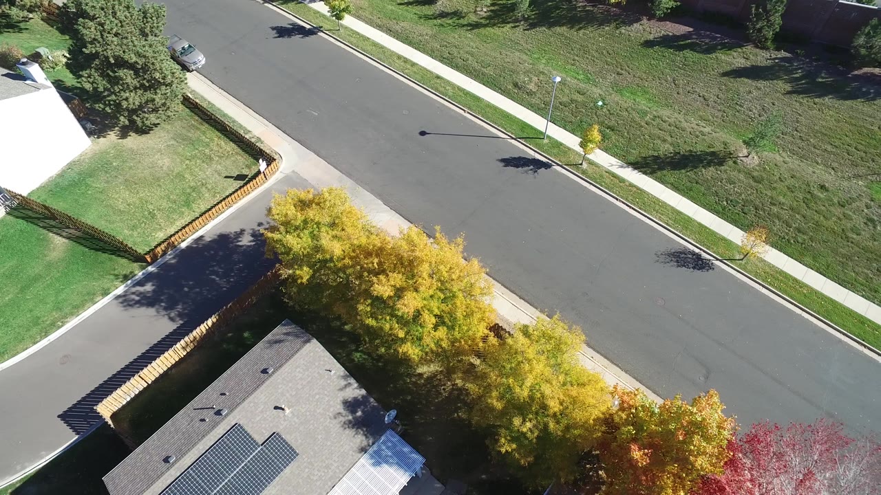 Overhead Hidden Lake, Colorado frame with the road centered, a rooftop and yellow tree canopy at the bottom, grass lots and sidewalks across the street, and no visible people.