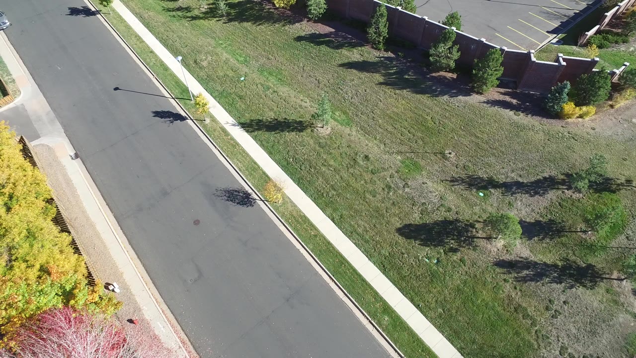 Opening aerial frame in Hidden Lake, Colorado showing a diagonal asphalt road, concrete sidewalk, grassy field, scattered trees, and part of a parking lot, with no visible people.