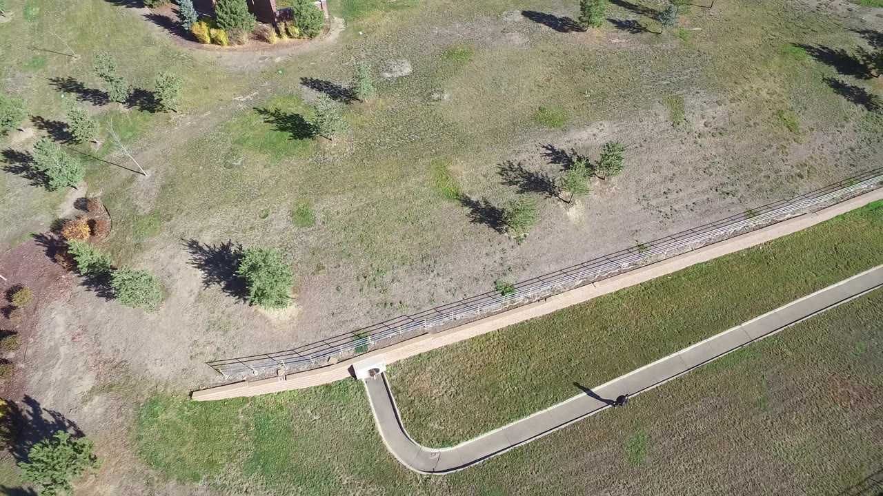 Aerial frame from Hidden Lake, Colorado with a pale curved access path, fence rail, dry grass, several trees, and no visible people.