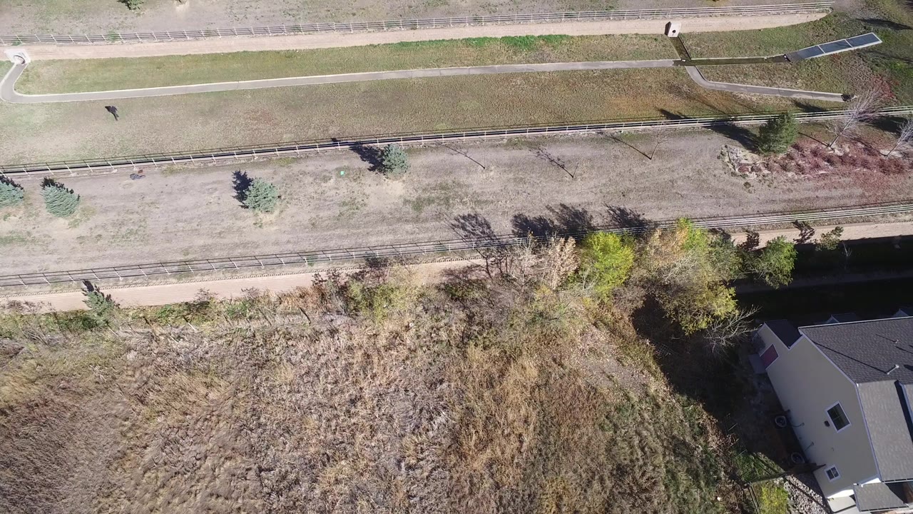 Overhead frame in Hidden Lake, Colorado showing a dry grass lot, paths, scattered trees, and the edge of modern homes, with no visible people.