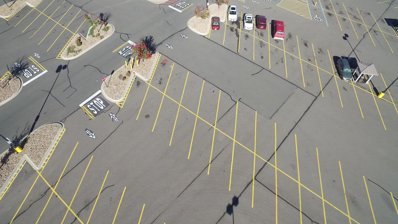 High-angle aerial view of a mostly empty parking lot in Hidden Lake, Colorado, with diagonal yellow parking lines, painted stop markings, landscaped islands, sparse cars at the top edge, and no visible people.
