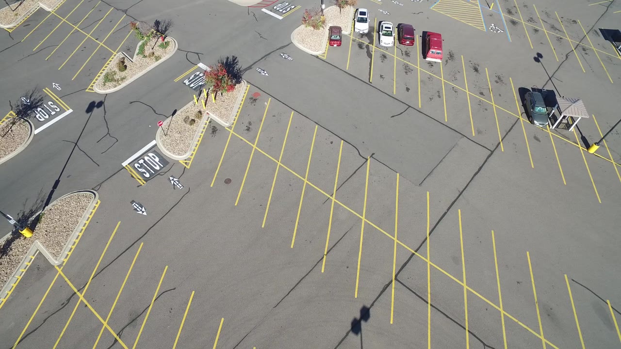 Overhead frame in Hidden Lake, Colorado, showing diagonal yellow parking stalls, a few parked cars near the upper edge, landscaped islands, light-pole shadows, and no visible people.
