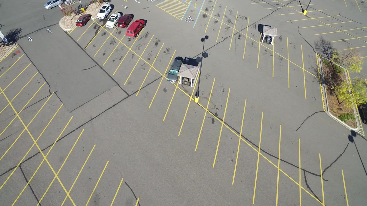 High aerial view over an almost empty parking lot in Hidden Lake, Colorado, with yellow stall lines, a few cars near the top edge, lampposts, asphalt cracks, and no visible people.