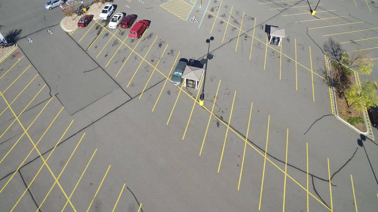 Drone-height view of the Hidden Lake parking lot with diagonal yellow no-parking markings, scattered cars near the upper left, a small structure near center, lamppost shadows, and no visible people.