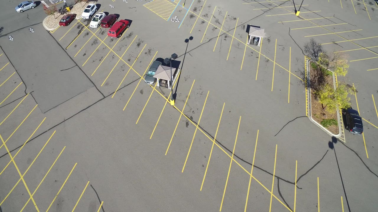 Mostly empty asphalt lot with yellow parking rows, lampposts, and a dark car near the right edge.