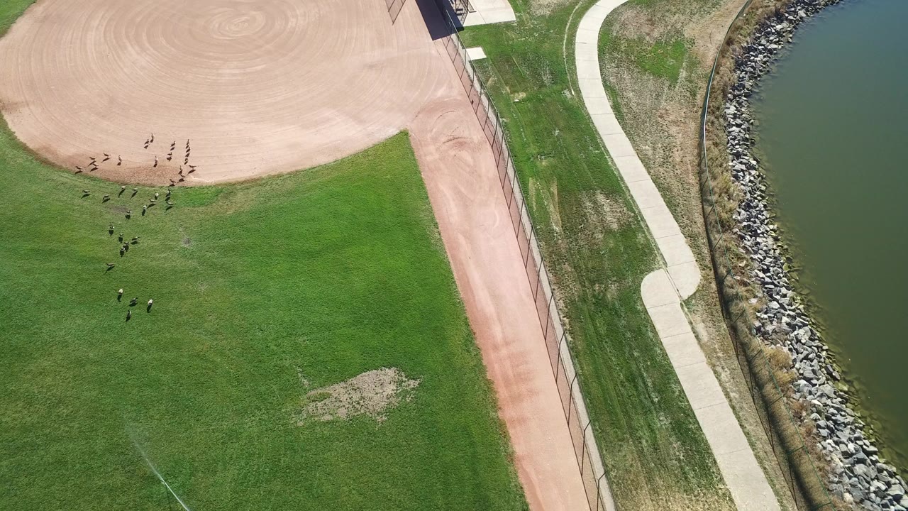 High aerial frame over Hidden Lake, Colorado showing green baseball outfield, tan infield dirt, a fence line, a curving path, rocky bank, green water, and no visible people.