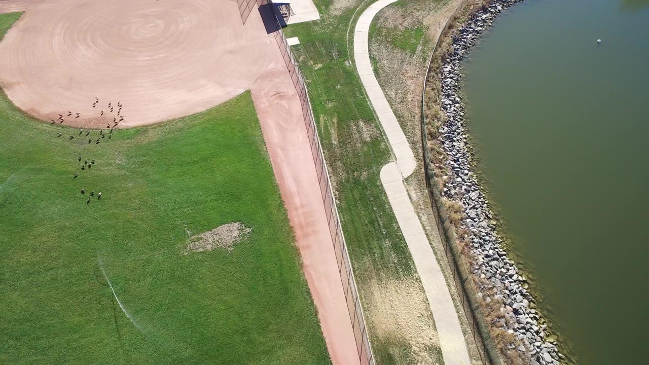 Overhead frame of Hidden Lake, Colorado with a green baseball outfield, tan infield edge, curved path, riprap shoreline, lake water, and no visible people.