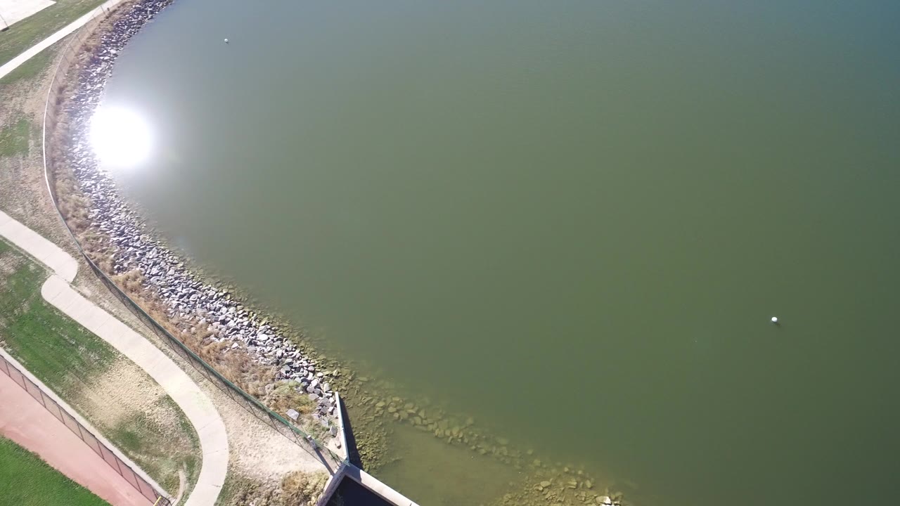 Overhead aerial frame from Hidden Lake Colorado showing green lake water, bright sun glare, a curved rock-lined shore, paths, a dark structure edge, and no visible people.