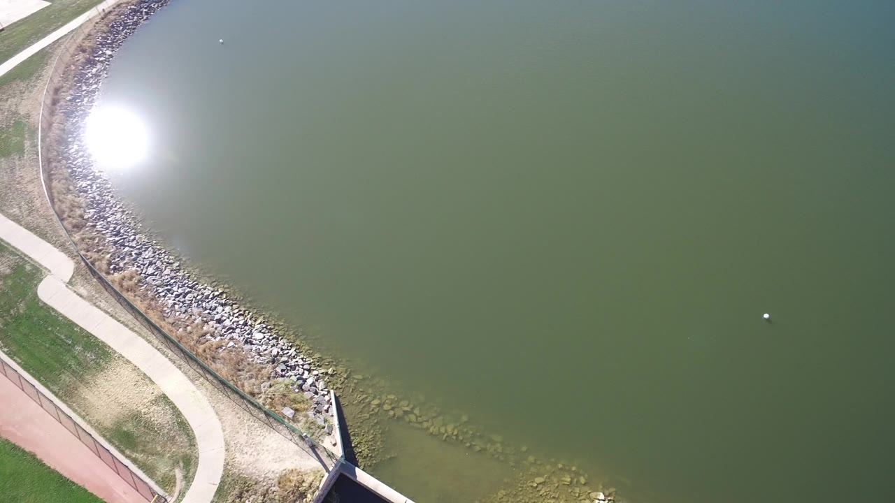 A high overhead view in Hidden Lake Colorado keeps the green water dominant while the sun reflection sits near the rock-armored shoreline, with no visible people.