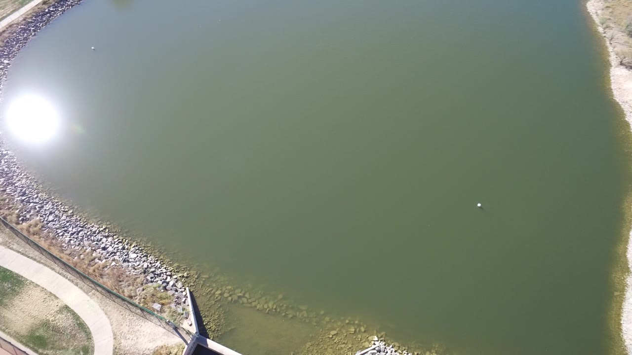 Drone view across green water with a rocky bank, path, and pale shore at the edge.