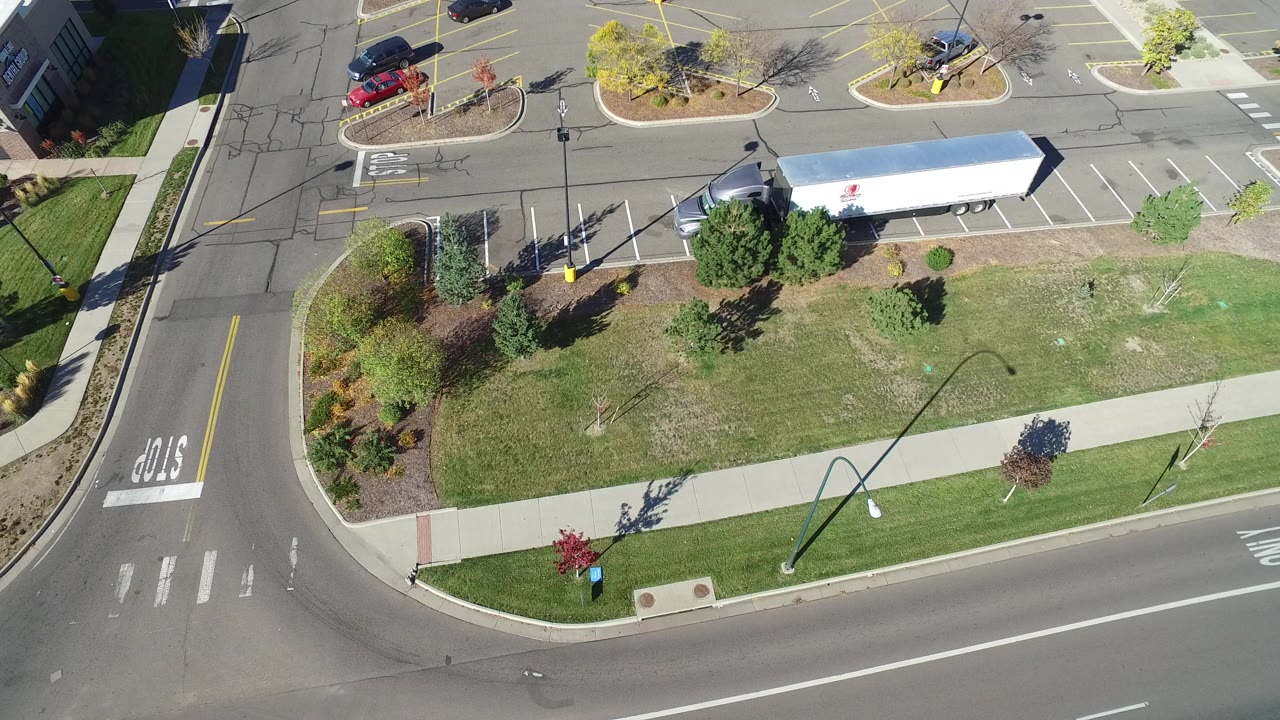 Overhead view of a road intersection, lawn, sidewalk, trees, parking lot, and white trailer.