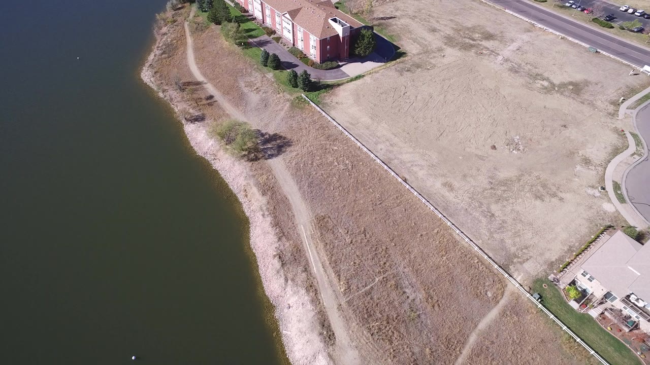 High oblique aerial frame over Hidden Lake, Colorado showing dark water on the left, a pale shoreline, dry grass, a fenced lot, nearby houses, a road, and no visible people.