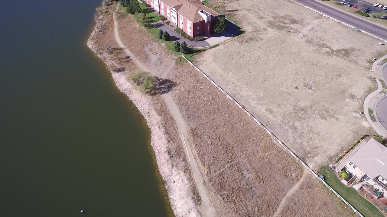 Aerial shoreline view with lake water, dry grass, a house corner, and road edge.