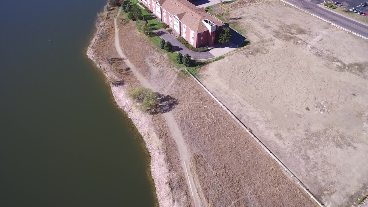 Aerial view of lake shoreline, dry grass, path, and red building.