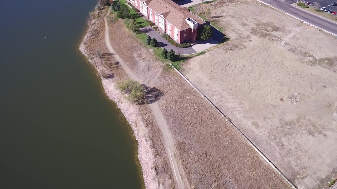 Hidden Lake, Colorado drone view with lake water on the left, a pale shoreline path below, dry grass through the center, and the red building set near the top edge with no visible people.