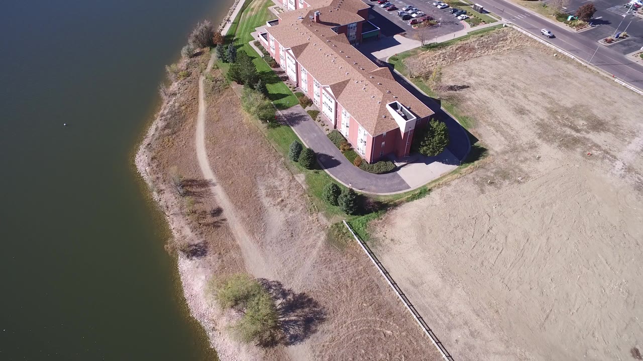 A high-angle aerial frame in Hidden Lake, Colorado shows a tan-roofed red residential building beside dark water, a pale shoreline path, dry open ground, and no visible people.