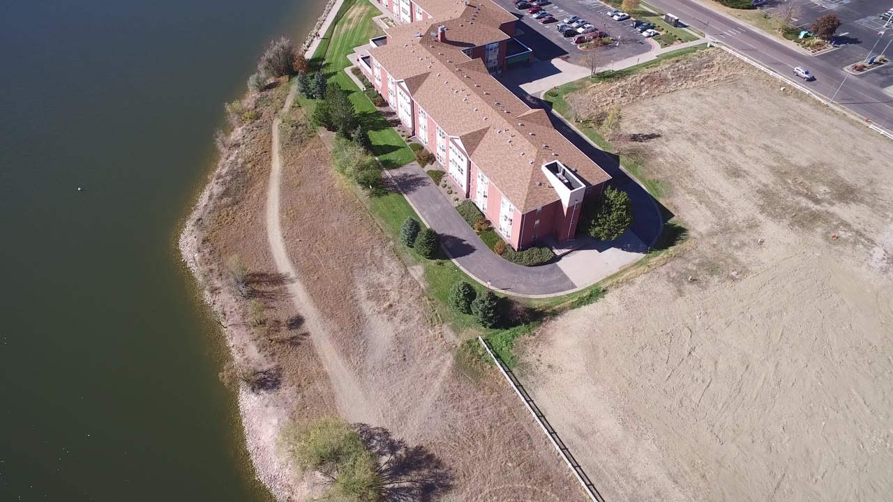 Aerial view of a tan-roofed building, lakeshore path, road, and parking lot.