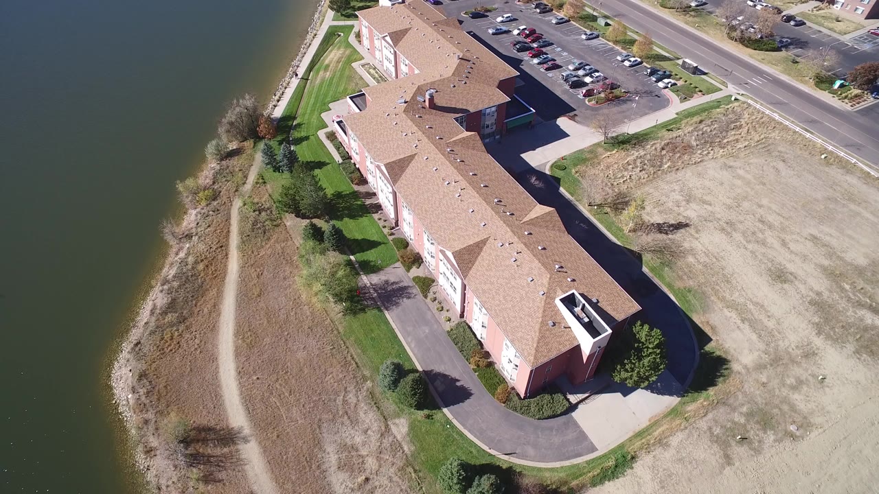 Aerial view of a long tan-roofed residential building beside lake water and a shoreline path.