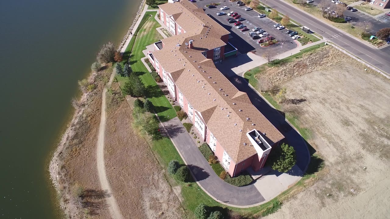 High aerial frame in Hidden Lake, Colorado with the long residential building running diagonally, lake water to the left, parked cars near the road, and no visible people.