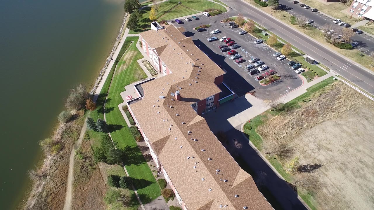 High overhead frame of Hidden Lake, Colorado showing a tan-roofed building, green lake water, parked cars, road, grass, and dry ground with no visible people.