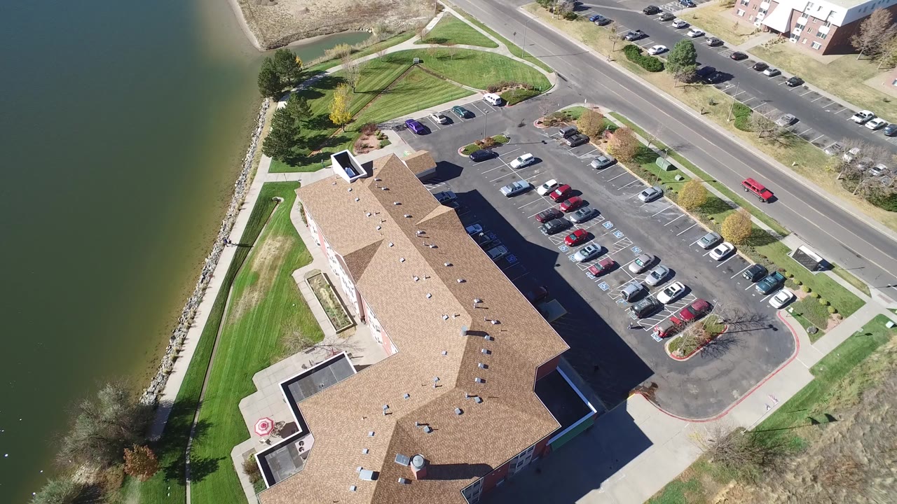 High aerial frame over Hidden Lake, Colorado showing green lake water beside a rock shoreline, a tan-roofed building, parked cars, a road, and no visible people.