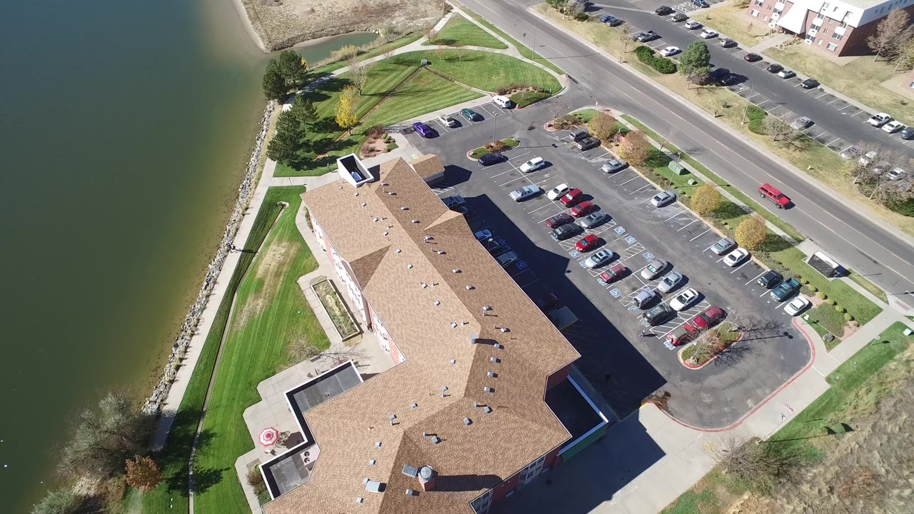 Second aerial frame over Hidden Lake, Colorado with the tan-roofed building centered lower in frame, cars lined in the lot, water to the left, and no visible people.