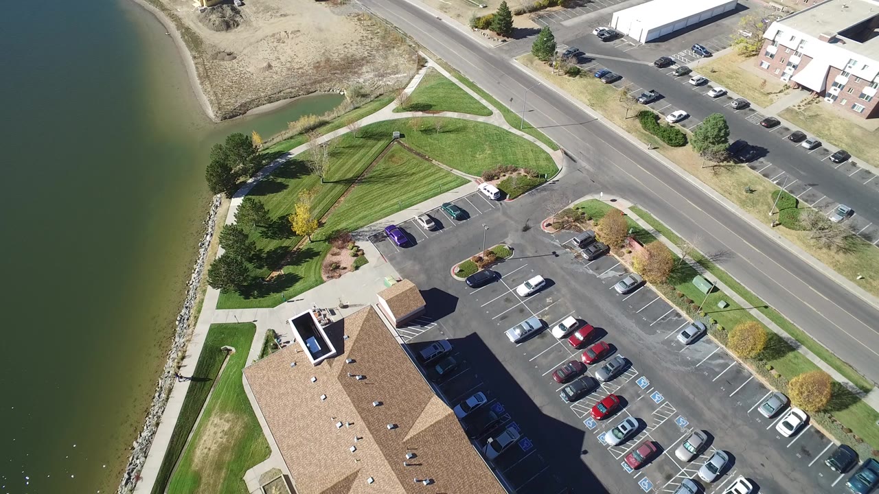 Aerial view of lake edge, parking lot, road, lawns, and low buildings.
