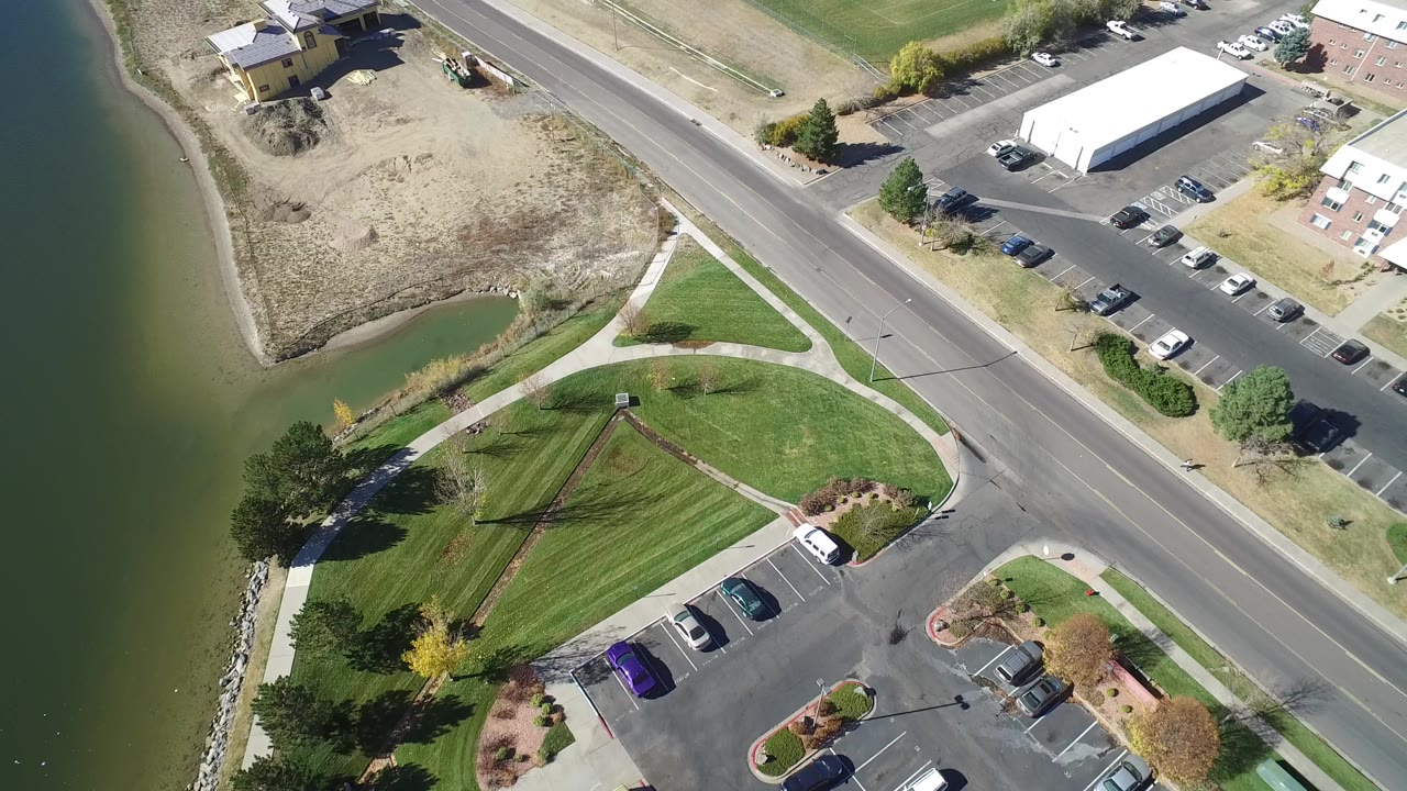 Aerial view of lake edge, park paths, road, parking lot, and white building.