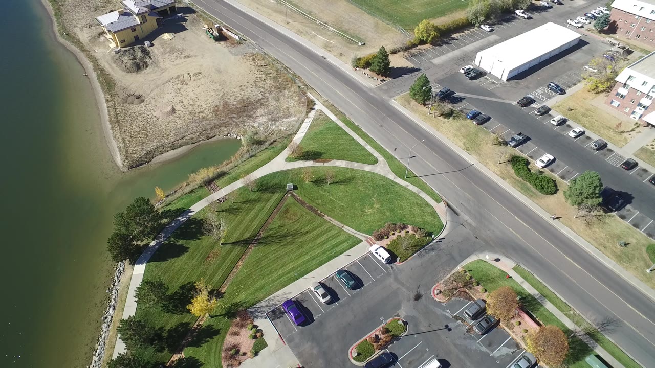 High-angle Hidden Lake, Colorado view with water on the left, triangular park paths below, a road crossing the upper frame, parked vehicles, and a white-roofed building with no visible people.