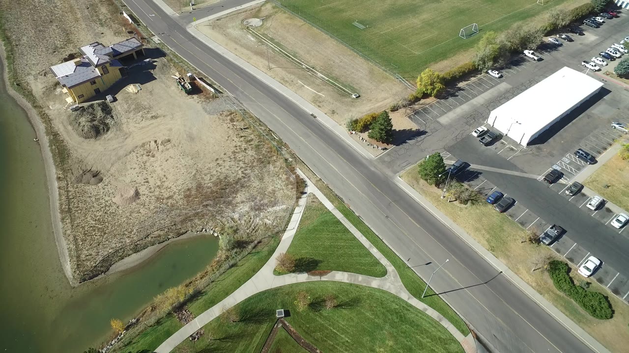 High overhead view in Hidden Lake, Colorado with a white-roofed business building, parked cars, a road, dry land, green sports fields, and no visible people.