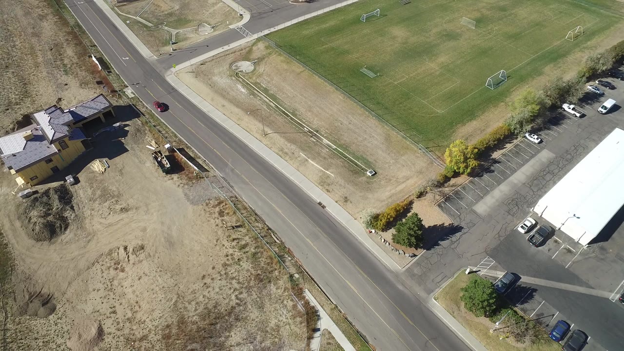 High overhead frame in Hidden Lake, Colorado showing a diagonal paved road, a green field with goal frames, a small parking lot, dry lots, rooftops, and no visible people.