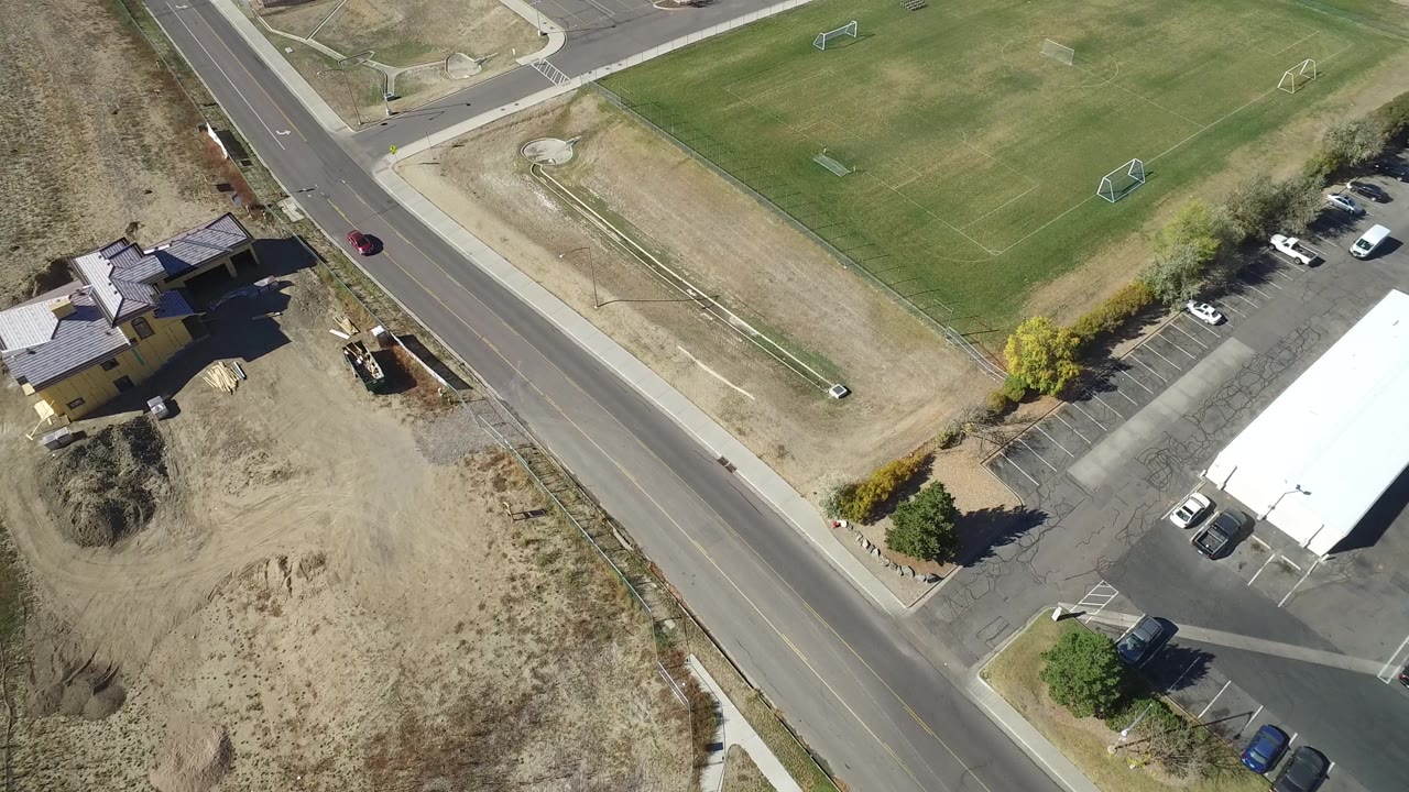 Overhead Hidden Lake, Colorado view with the sports field taking more of the upper frame, a road crossing diagonally, parked vehicles by a white-roofed building, homes, and no visible people.