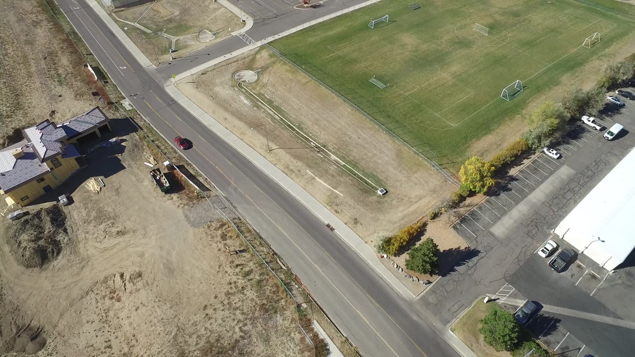 Top-down view of a road beside a sports field, parking spaces, dry land, and homes.