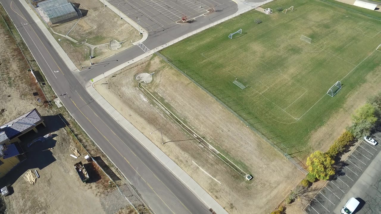 Second overhead frame in Hidden Lake, Colorado with the road running along the left edge, soccer goals on the field, tan undeveloped ground, parking pavement, and no visible people.
