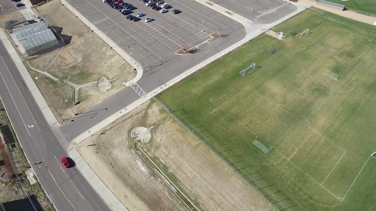 High overhead frame in Hidden Lake, Colorado showing a paved parking lot, a road, sidewalks, and green soccer fields with no visible people.