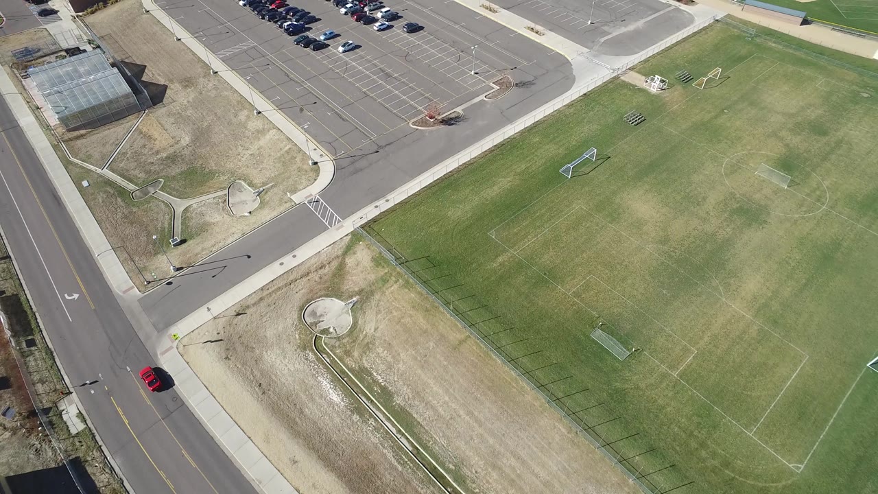 Bird's-eye view in Hidden Lake, Colorado with rows of parked cars at the top, road pavement at left, and marked soccer fields filling the right side, with no visible people.
