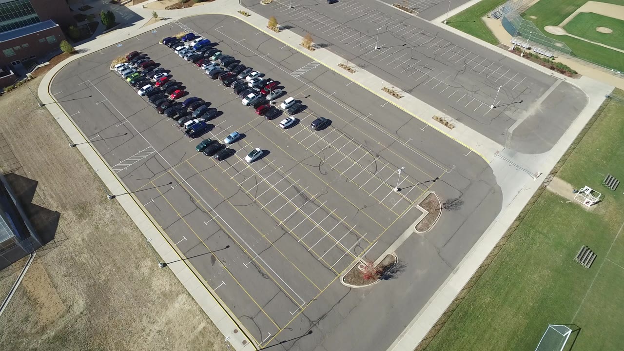 Aerial frame in Hidden Lake, Colorado showing parked cars grouped near the left side of a large asphalt lot, with curving access roads, lawns, and no visible people.