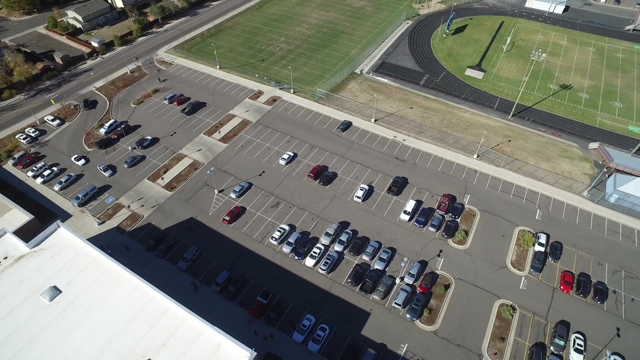 Top-down aerial frame in Hidden Lake, Colorado showing a large parking lot, parked cars, a white-roofed building edge, a road, and a green athletic field with no visible people.