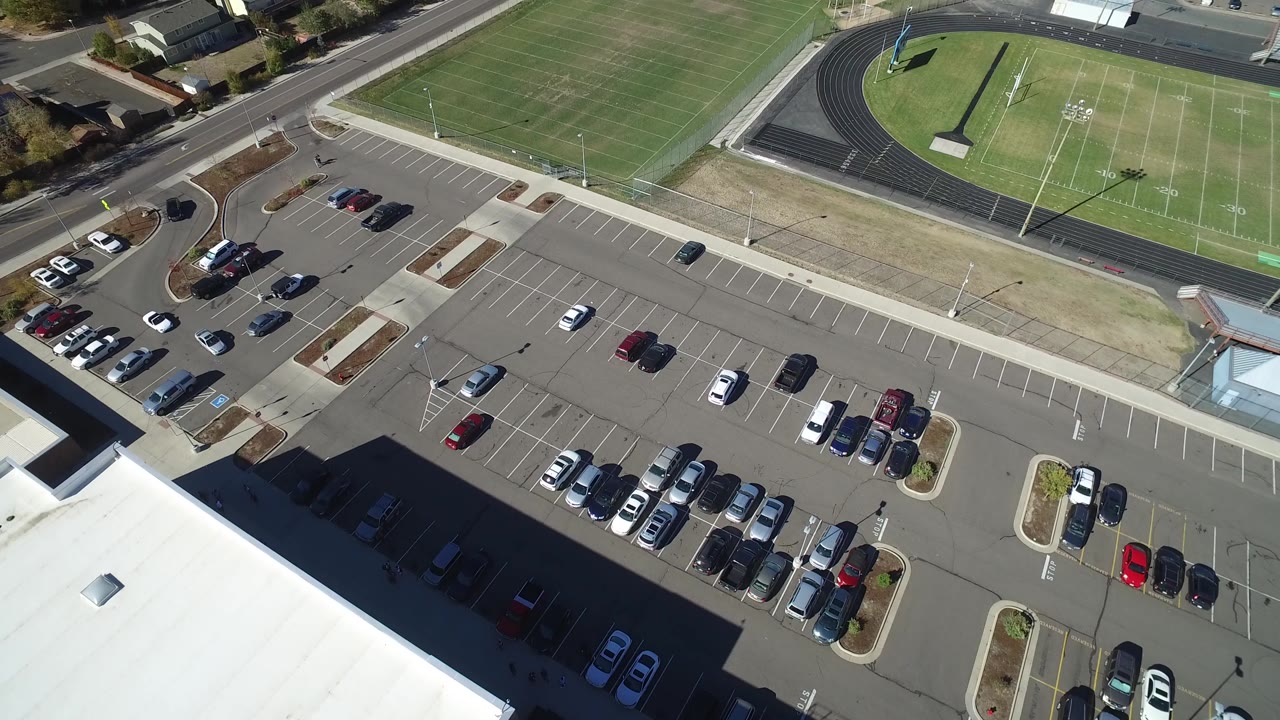 High aerial view in Hidden Lake, Colorado with rows of parked cars, empty parking spaces, a roadway, and the sports field and track extending across the top of the frame, with no visible people.