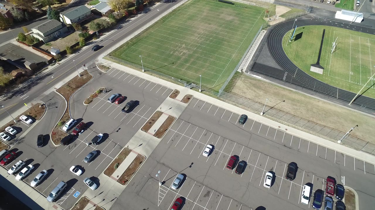Overhead frame in Hidden Lake, Colorado showing a green football field beside a black running track, a mostly empty parking lot below, bordering roads and homes, and no visible people.