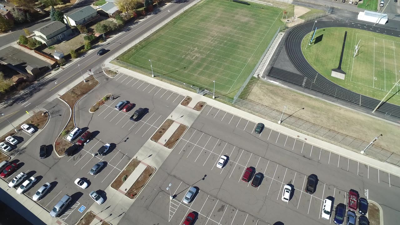 Aerial frame in Hidden Lake, Colorado with the football field across the top, the track oval at right, scattered cars in the parking lot below, residential roofs along the road, and no visible people.