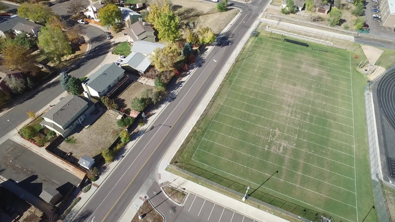 Aerial frame from Hidden Lake, Colorado with the football field shifted across the right side, a road running vertically near center, private homes to the left, and no visible people.