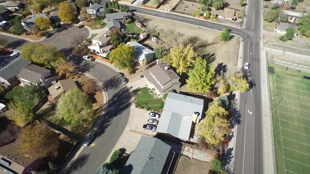 Opening aerial frame over Hidden Lake, Colorado shows residential roofs, driveways with parked cars, lawns, autumn trees, a curved street, and no visible people.
