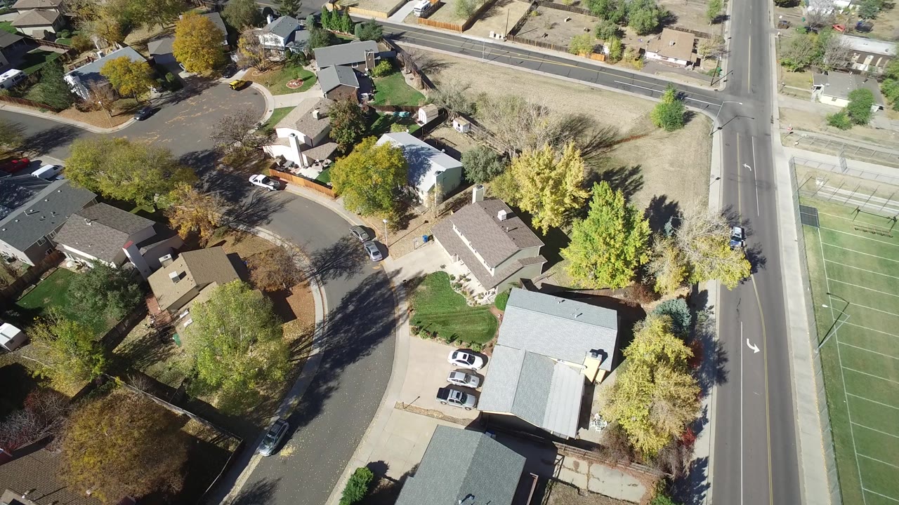 Overhead neighborhood view with houses, yellow trees, driveways, parked cars, and roads.