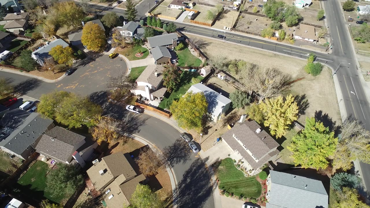 High overhead view in Hidden Lake, Colorado with homes clustered around a curved street, a white vehicle by a driveway, fenced yards, green and yellow trees, and the straight road along the upper edge.