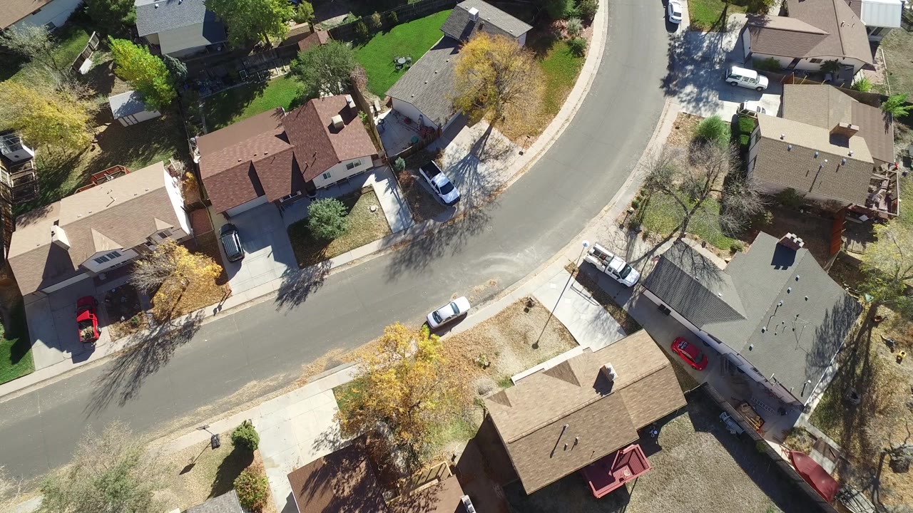 High aerial frame in Hidden Lake, Colorado with the road sweeping through the center, brown roofs and driveways on both sides, parked vehicles, mixed green and yellow trees, and no visible people.